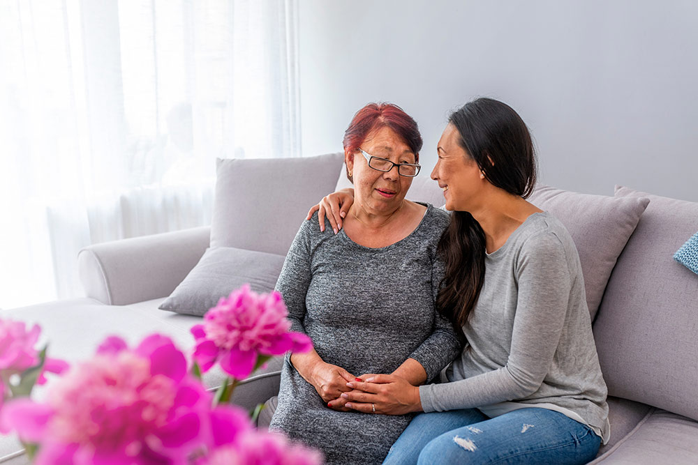 Senior woman sitting on couch with daughter, pink flowers in foreground