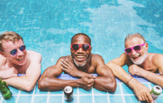 Three senior men smiling and laughing in pool with drinks and sunglasses