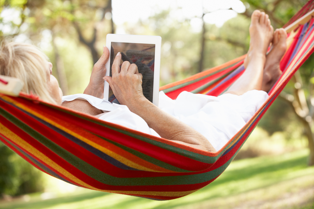 A senior woman lounges in a hammock while reading on her tablet